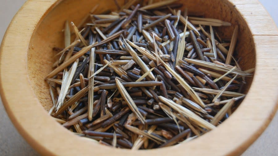 Several grains of manoomin, both light brown and dark brown, sit inside a small, wooden bowl.