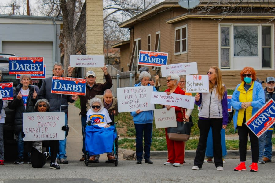 Protestors stand alongside the street holding signs stating "No tax payer funded charter schools", "no vouchers with tax dollars", and "no school vouchers".