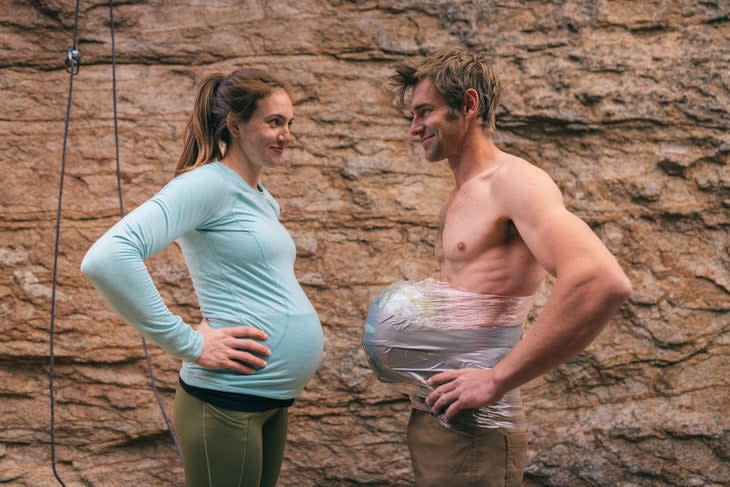 Pregnant woman in blue shirt faces shirtless man with watermelon taped to his stomach.