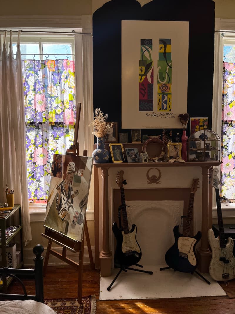Floral curtain hangs on window behind easel in combo office, guest room, studio, with guitars in front of mantel.