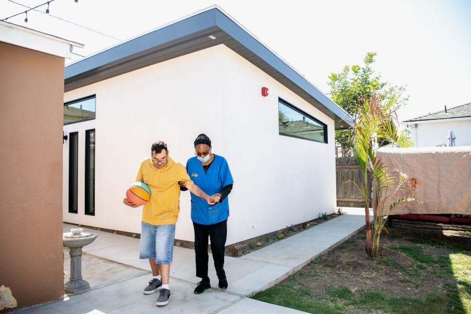 A man carrying a basketball walks with his caregiver outside a home.