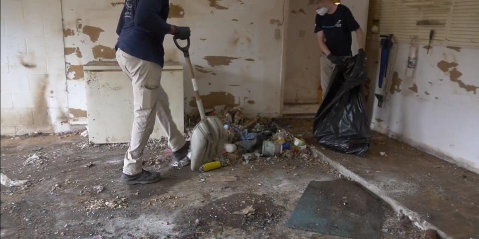 Two people clearing out trash and old flooring in a garage. One has a trash bag and the other has a shovel. Both are wearing light-colored pants and navy collared shirts.