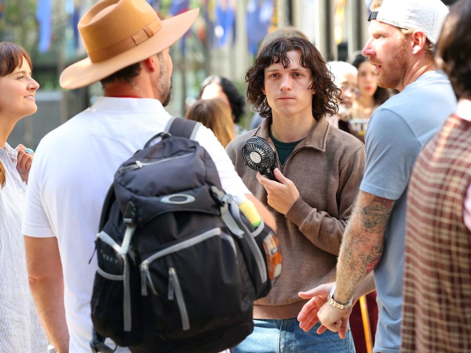 tom holland, wearing a brown sweater and jeans, holds a black fan and talks to seeral people while outside