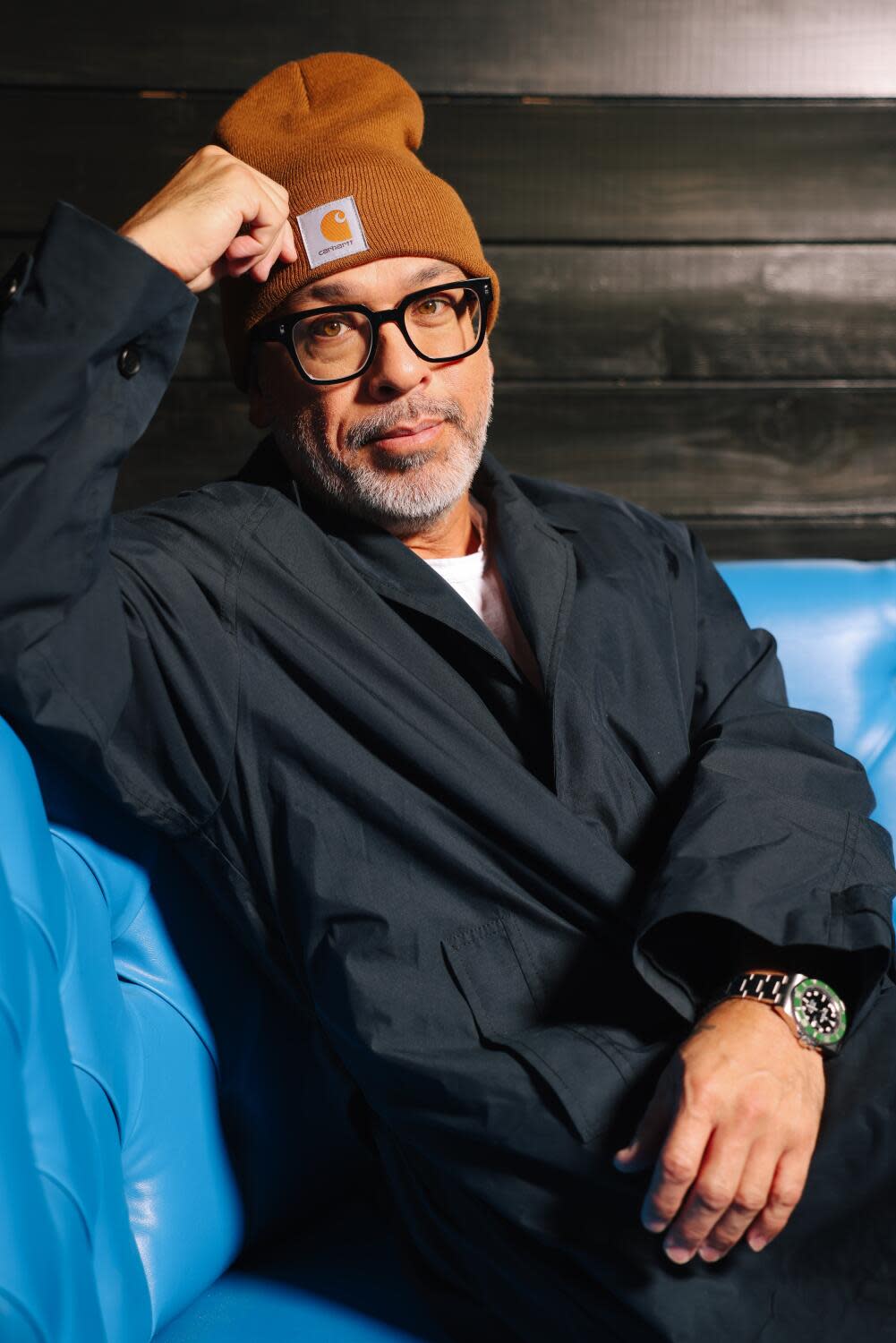 A mean in a beanie sits on a blue couch for a portrait.