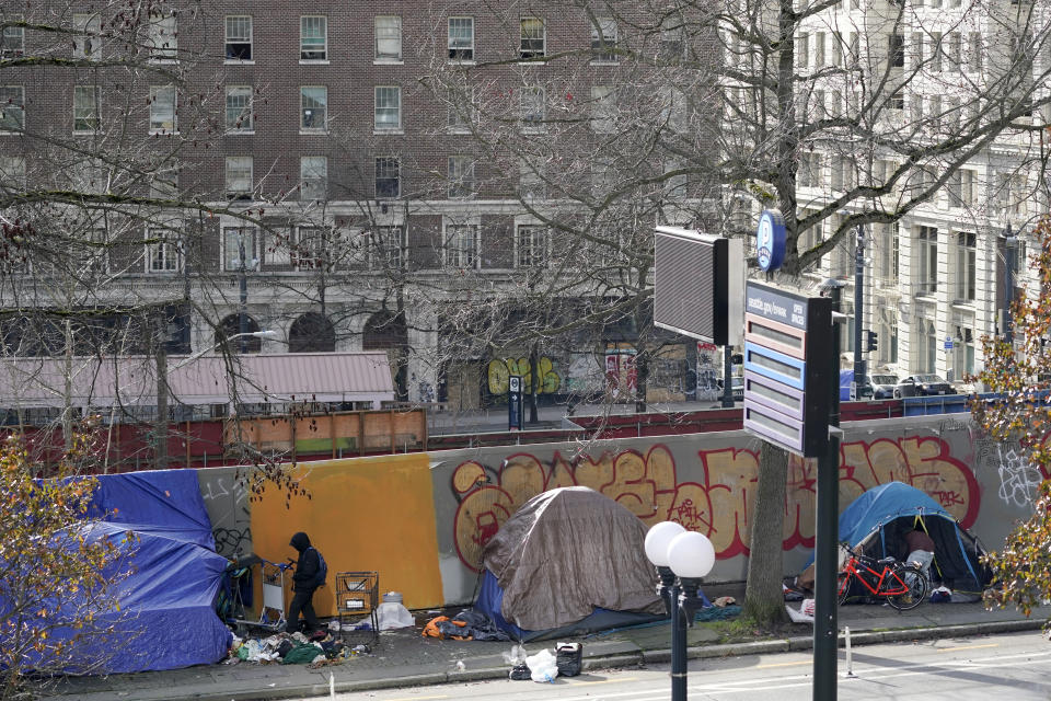 A person at left walks toward the entrance of a tent used by people experiencing homelessness, Tuesday, March 1, 2022, in downtown Seattle across the street from City Hall. For years, liberal cities in the U.S have tolerated people living in tents in parks and public spaces, but increasingly leaders in places like Portland, Oregon, New York and Seattle are removing encampments and pushing other strict measures that would've been unheard of a few years ago. (AP Photo/Ted S. Warren)