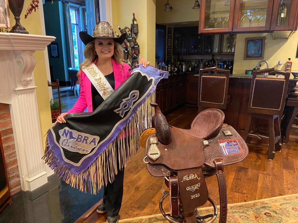 Mary Ivey, the National Little Britches Rodeo Queen for 2022-2023 stands in her living room with her pant chaps and brand new saddle from the association.