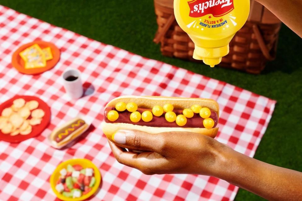 A mustard bottle held over a hot dog with mustard skittles near a picnic table with a red and white tablecloth