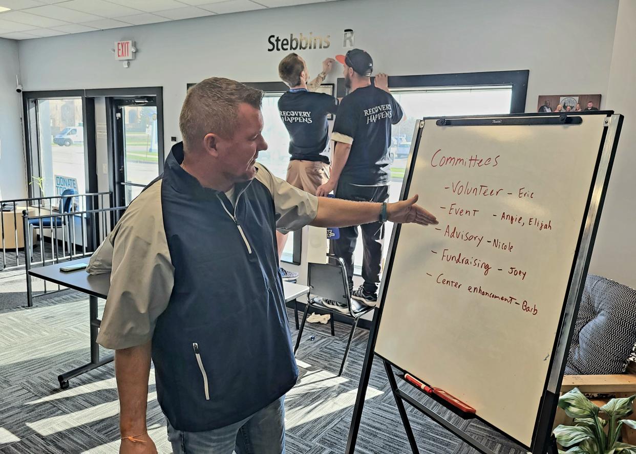 Patrick Patterson, director of Blue Water Recovery and Outreach Center, shows a breakdown of new committees on Friday, April 14, 2023, while volunteers etch the name of a supporting foundation on the wall nearby at their 10th Street facility in Port Huron.
