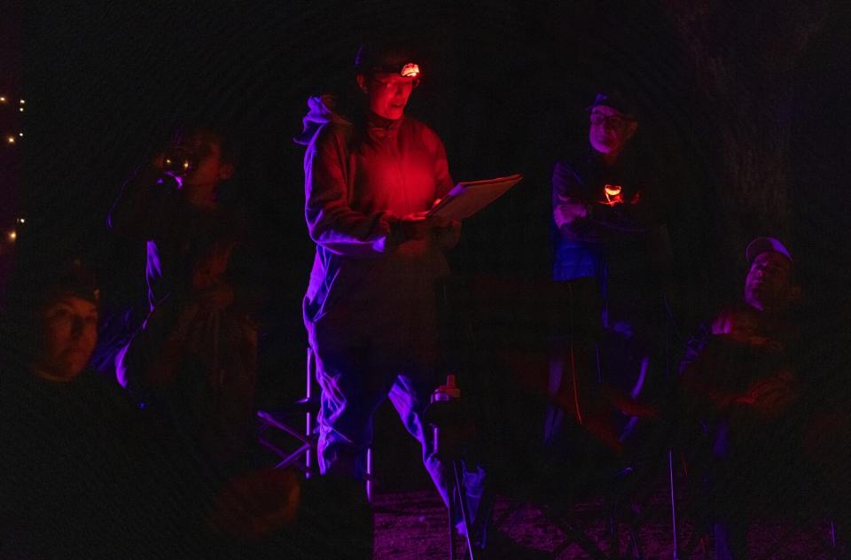 Dani Hsia, center, a headlamp illuminating her handwritten notes, speaks to a group sitting around a campfire.