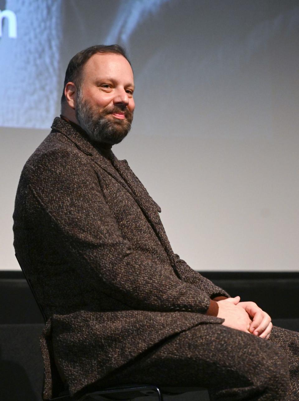 yorgos lanthimos sits on a stage and smiles out toward the audience, he wears a brown suit, behind him is a projection on a large screen