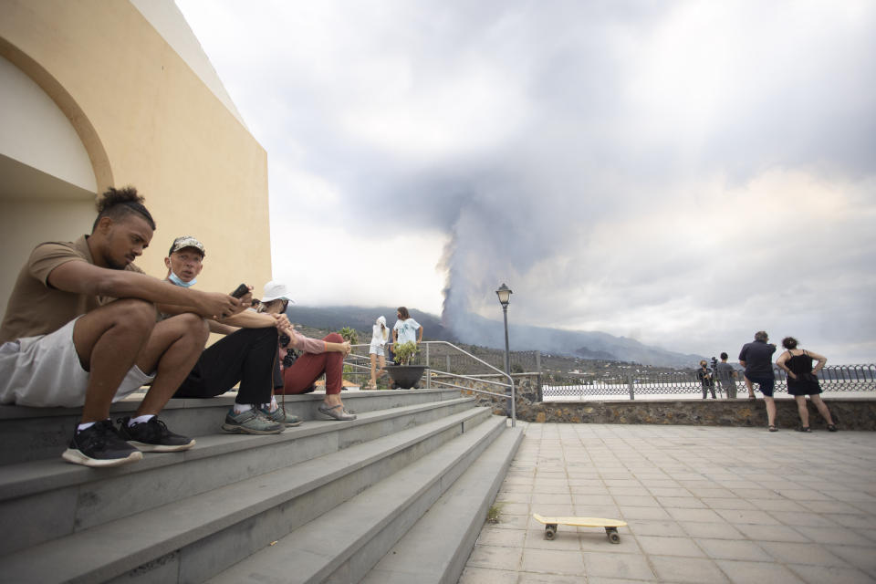 People sit on some steps as others look at the eruption of a volcano near El Paso on the island of La Palma in the Canaries, Spain, Monday, Sept. 20, 2021. Lava continues to flow slowly from a volcano that erupted in Spain's Canary Islands off northwest Africa. Officials say they are not expecting any other eruption and no lives are currently in danger. (AP Photo/Gerardo Ojeda)