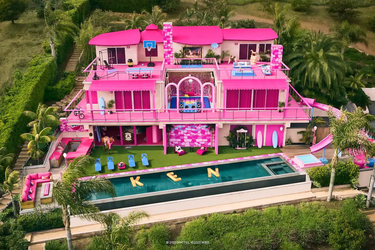 An exterior of the Barbie Dreamhouse on Airbnb shows the famously pink house surrounded by amenities such as a pool, decks, workout equipment, and more.