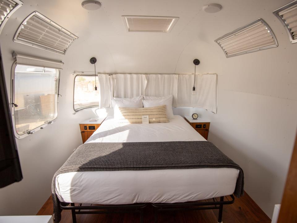 A bedroom inside Autocamp Joshua Tree's Airsteram trailer with white accents, a bed with pillows, curtains over windows.