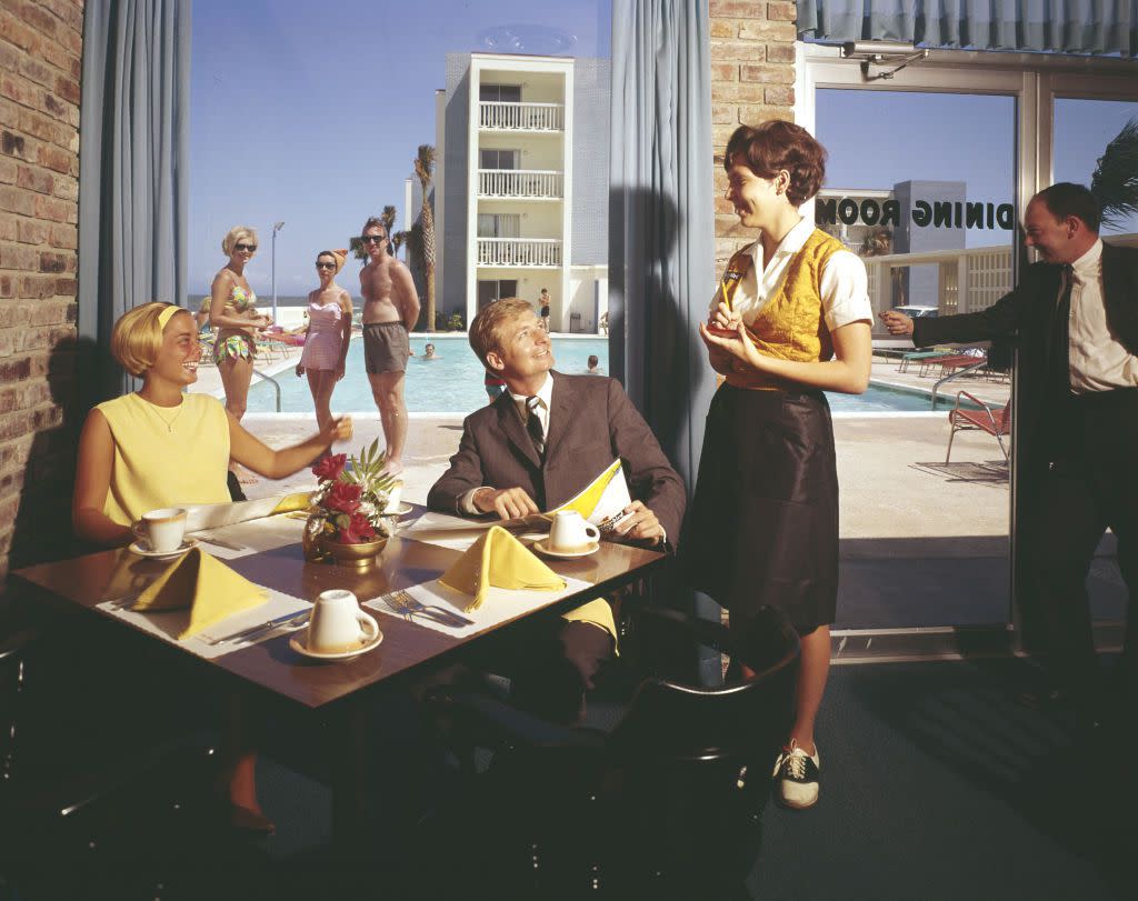 While several people in swimwear watch from outside the floor-to-ceiling windows, a smiling couple place an order with a smiling, uniformed waitress in the coffee shop of the Thunderbird Motel, Myrtle Beach, South Carolina, 1960s