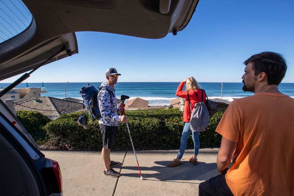 A man with a cane stands with another man and a woman near the ocean.