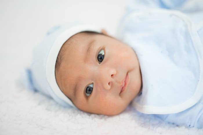 A close-up of a baby lying on a soft surface, wearing a hat and swaddled. The baby has large, curious eyes and a calm expression