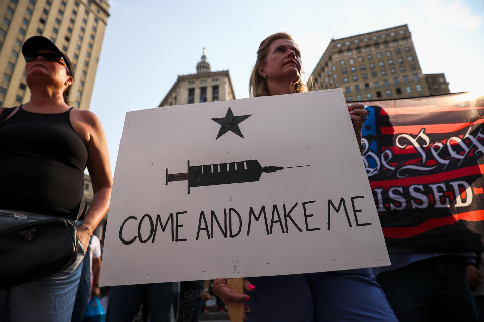 Protesters stand against a backdrop of skyscrapers holding signs, one of which shows an image of a hypodermic needle and reads: Come and make me.