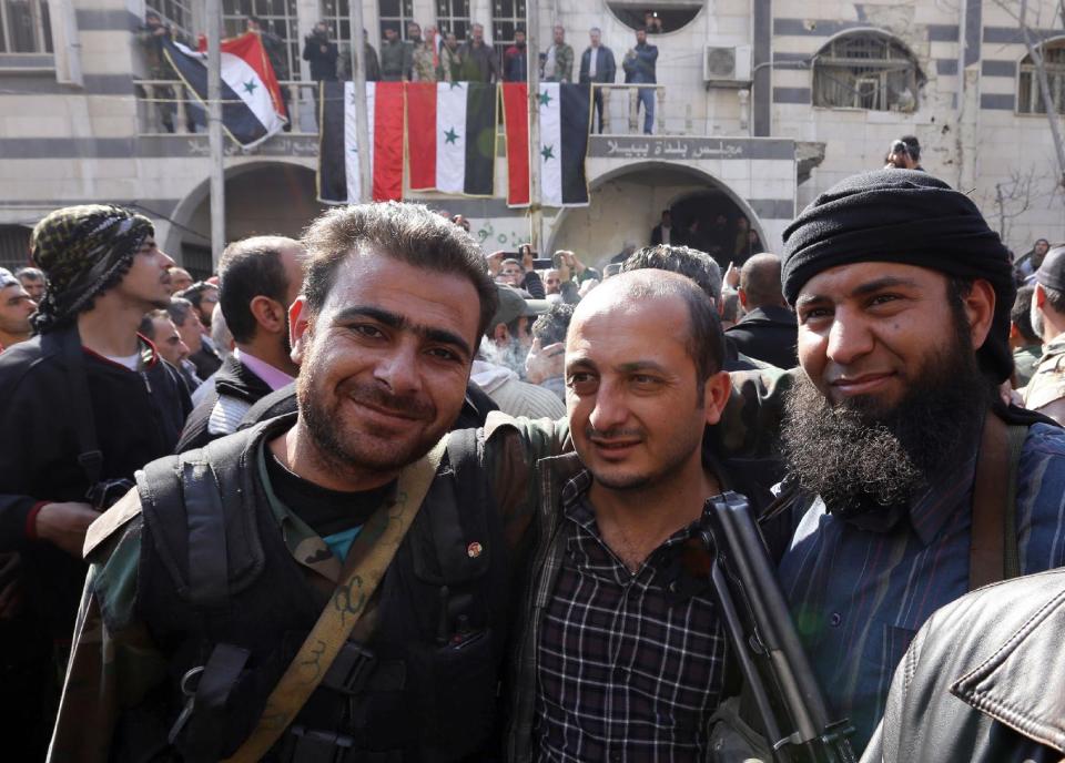 In this Monday Feb. 17, 2014 photo, Syrian rebels, right, stands with two soldiers from Syrian government forces, center and left, as they pose for a picture after a reconciliation agreement between the two groups, in Babila, southeast Damascus, Syria. In one besieged neighborhood after another, weary rebels have turned over their weapons to the Syrian government in exchange for an easing of suffocating blockades that have prevented food, medicine and other staples from reaching civilians trapped inside. The government touts the truces as part of its program of “national reconciliation” to end Syria’s crisis, which has killed more than 140,000 people since March 2011. (AP Photo)