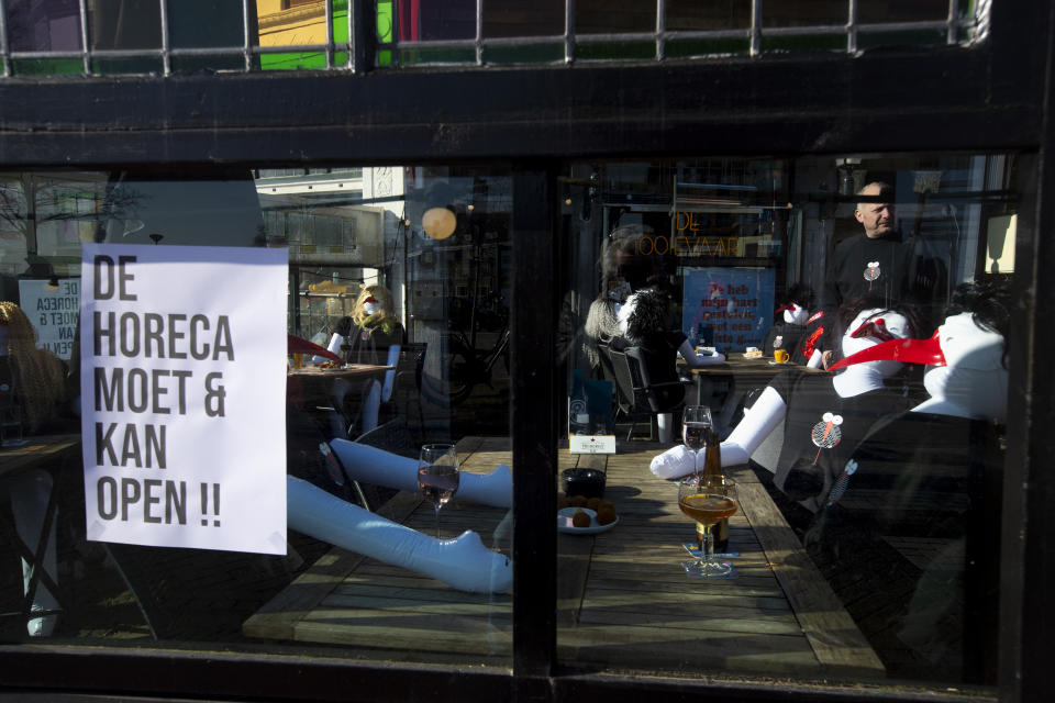 A sign reads "Hotel, Restaurants, Cafes, can and must open", as owner Peter Bender, rear right, of cafe De Ooievaar talks to a passer-by on his terrace filled with inflatable dolls at cafe in Delfshaven, Rotterdam, Netherlands, Tuesday, March 2, 2021. Stores in one village opened briefly, cafe owners across the Netherlands were putting tables and chairs on their outdoor terraces and sex workers were planning a demonstration outside parliament in protests against the government's tough coronavirus lockdown. (AP Photo/Peter Dejong)