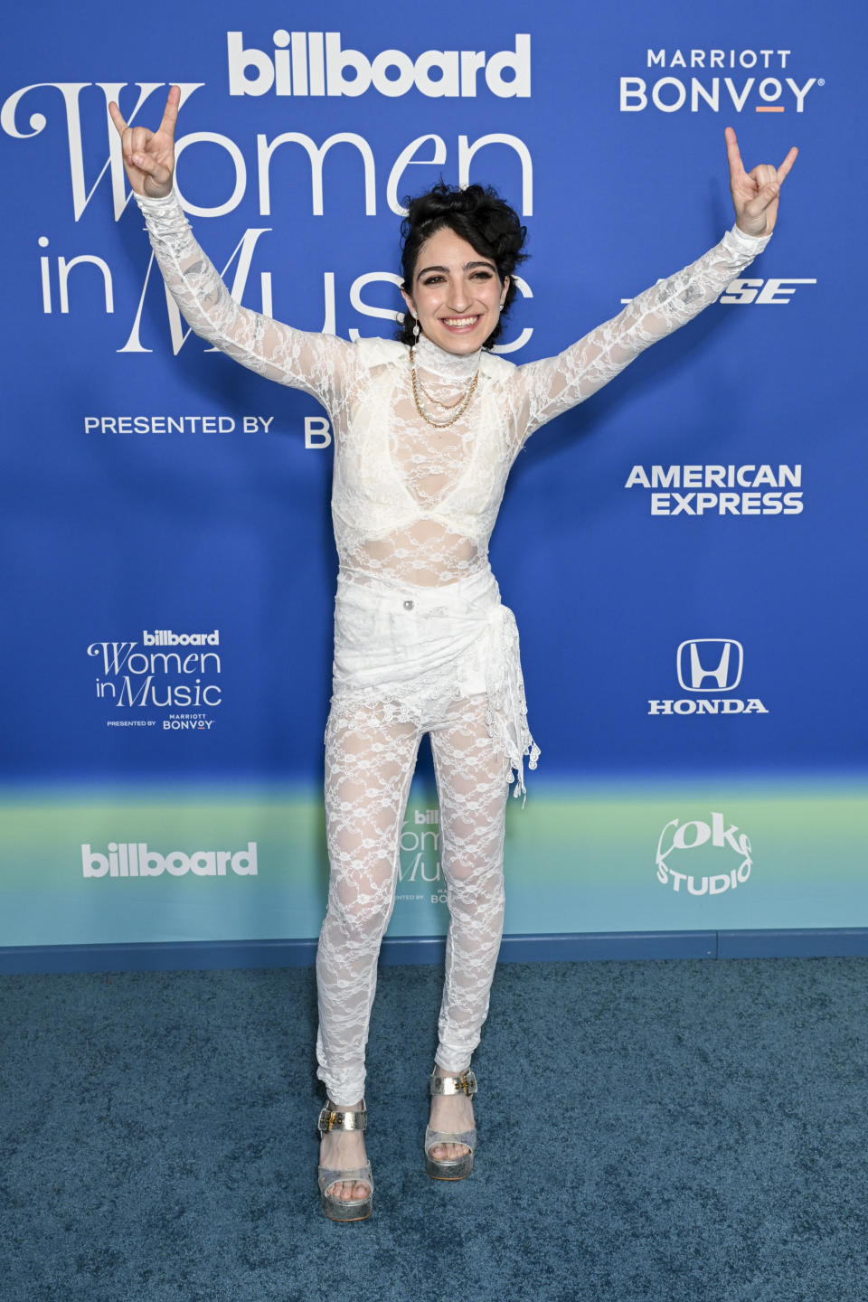 Emily in lace sheer jumpsuit over a bralette and shorts with hands up