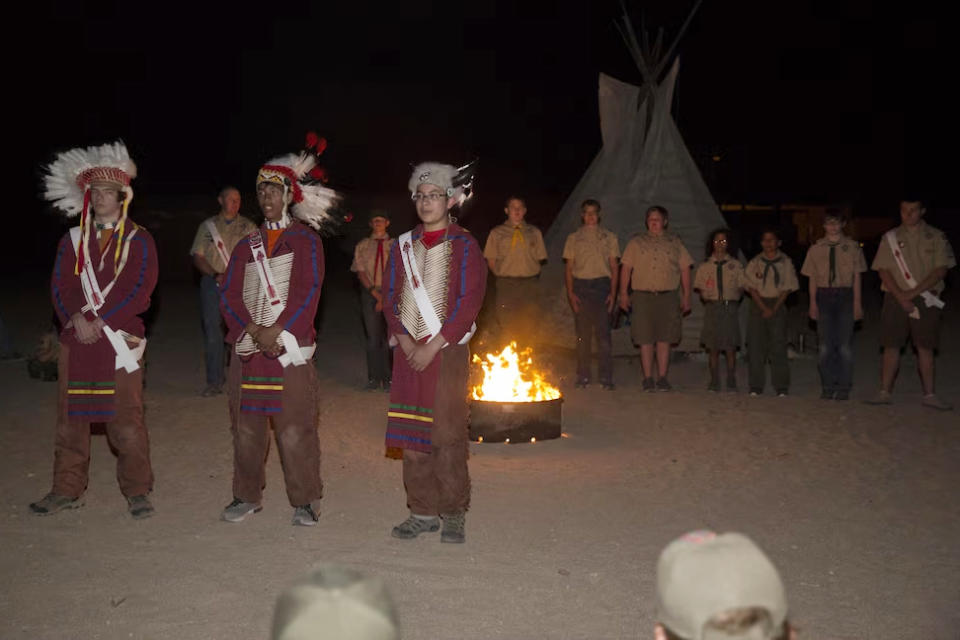 Order of the Arrow nominees stand in line, background, as three members of the order, dressed in Native American regalia, explain what the nomination of the scouts behind them signifies during their Call Out Ceremony at Camp Wilson during the Boy Scout Camp Out for local Boy Scouts of America troops in 2016. (Sgt. Charles Santamaria / U.S. Marines)