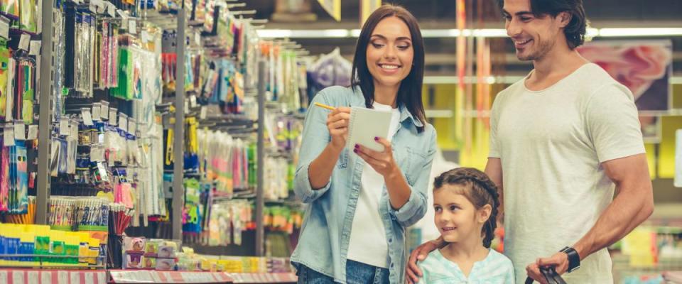 Beautiful young parents and their cute little daughter are smiling while choosing school stationery in the supermarket. Mom is making notes in the list