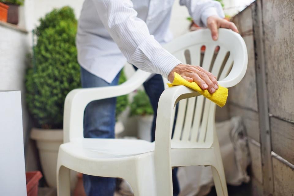 Person wearing a white shirt cleaning a white plastic chair on a patio with a yellow cloth.