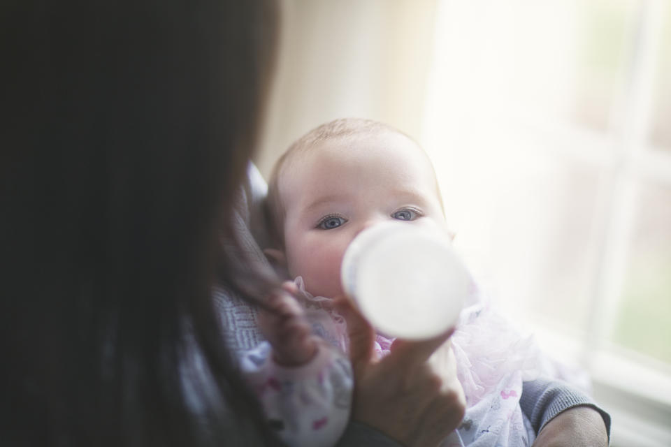 A baby is being fed with a bottle by an adult holding them. The baby looks towards the camera