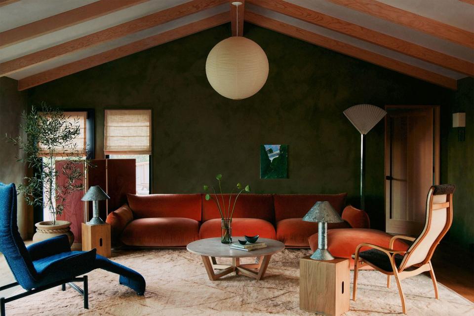 living area with peaked ceiling and a large globe pendant at center and an orange velvet three cushion sofa against a greenish wall and some box crate side tables with blue metal lamps and a fancy blue lounge chair on a crinkle style off white rug