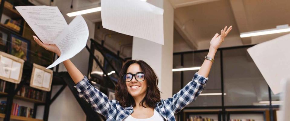 Joyful young brunette woman in black glasses throwing papers above in library. Cheerful mood, smiling to camera, good results, expressing positive true emotions