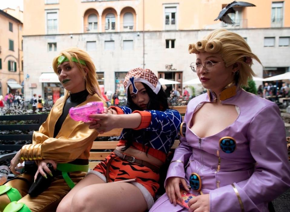 Three individuals in vibrant cosplay costumes from the anime JoJo's Bizarre Adventure sit on a bench, with one holding a toy gun and pointing it forward