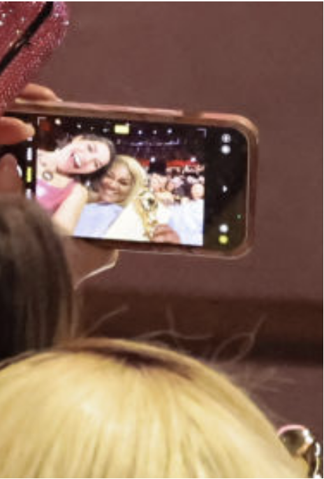 Person taking a selfie with two celebrities at an event, visible on smartphone screen
