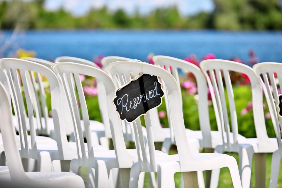 White plastic chairs arranged in rows at an outdoor wedding venue by a lake, with a "Reserved" sign on one chair