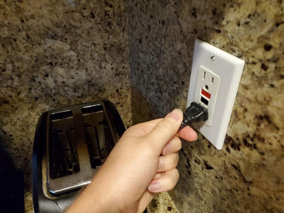 Close-up, personal perspective image of human hand of a man plugging an electrical cord into a Ground Fault Circuit Interrupter (GFCI) electrical outlet on the wall in a domestic room, September 12, 2019