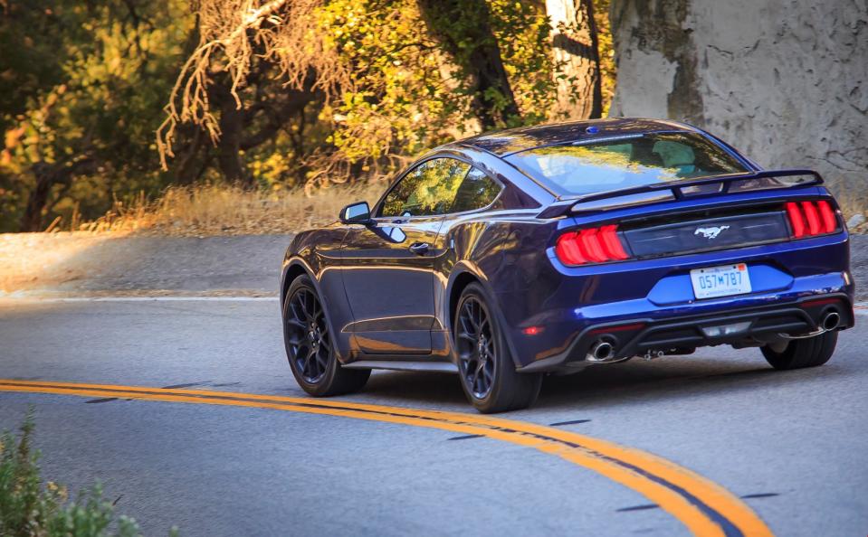 Blue Ford Mustang rounding a corner on gray asphalt with bushes along the roadside.