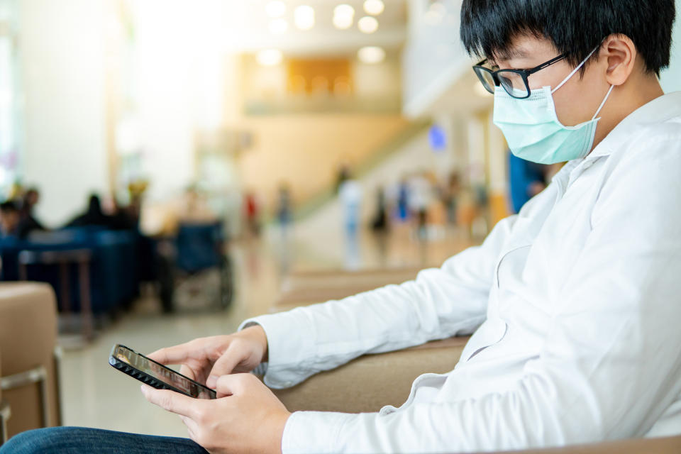 Male Asian patient wearing surgical mask using smartphone at waiting area in hospital or medical center. Medical exam or body check up. Wuhan coronavirus outbreak prevention. Health care concept