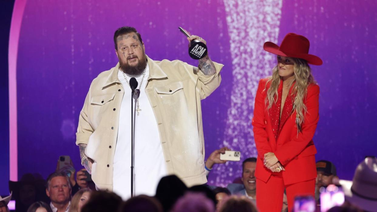 jelly roll holds a trophy in one hand and speaks into a microphone while standing on a stage, lainey wilson stands on the right and looks at him as fans watch from below the stage
