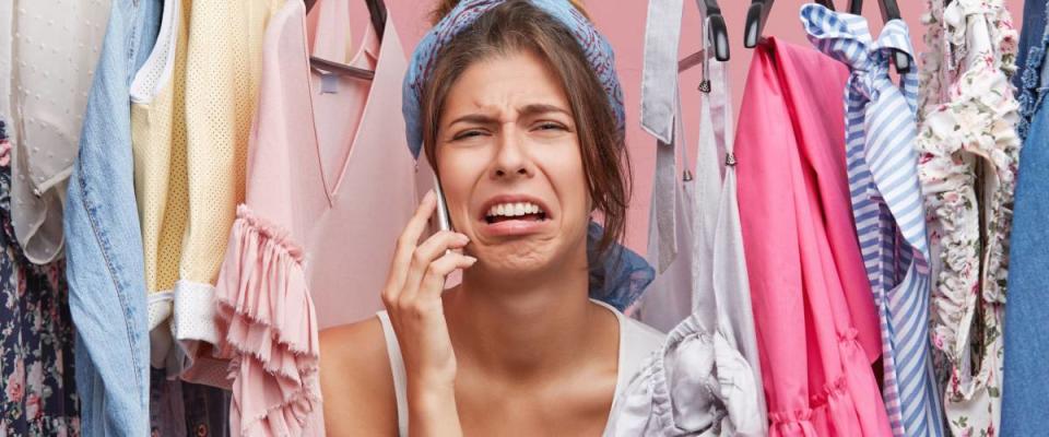 Upset woman standing near rack of clothes, chatting over smart phone with her friend, complaining that she has nothing to wear. Displeased female not knowing what to put on for birthday party