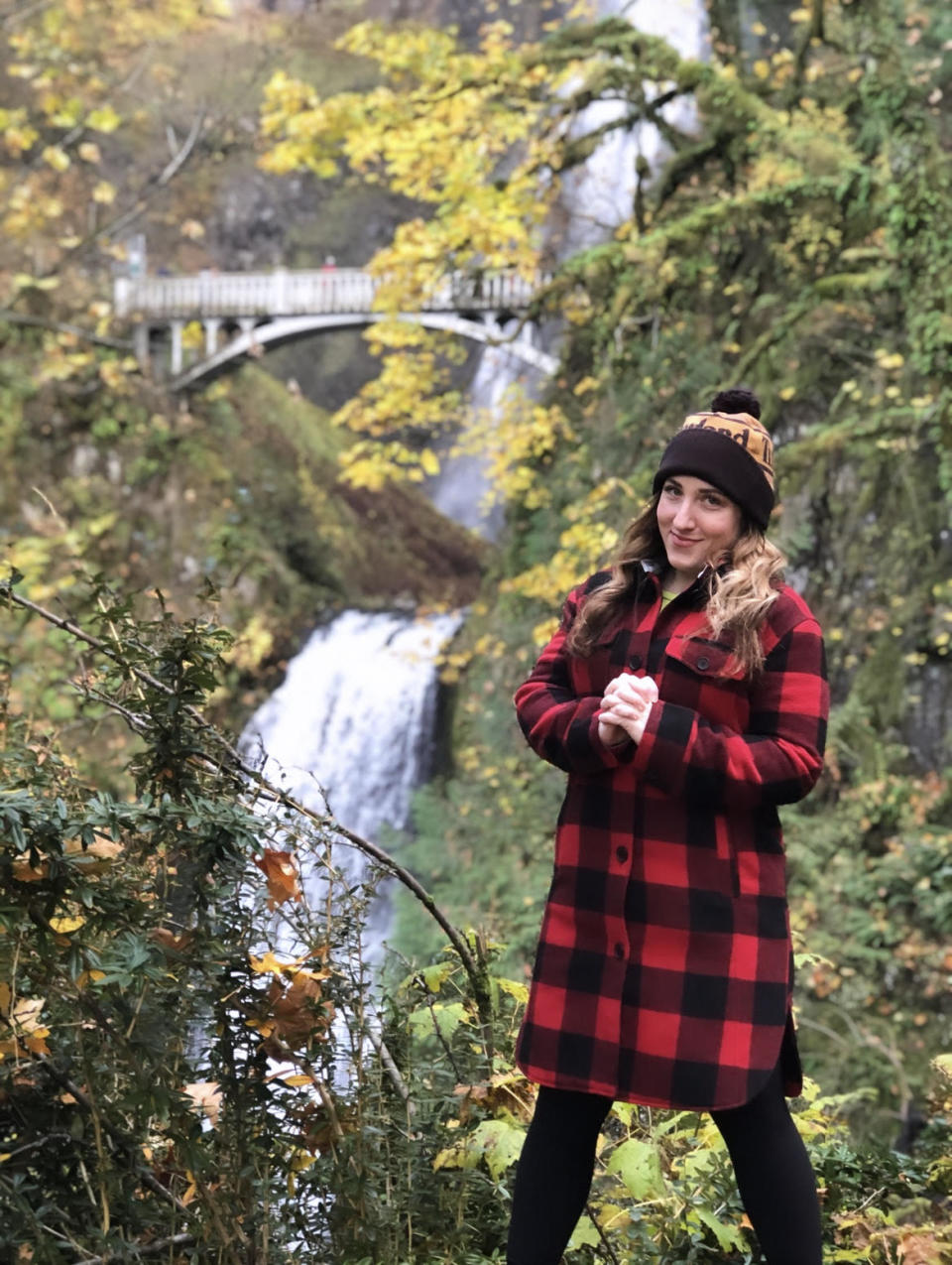 Mia, who had Bartholin's cysts, wears a red-and-black coat and a brown-and-beige hat standing in a forest with a waterfall behind her. 