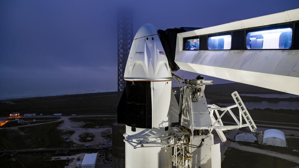 A black and white spacecraft on a white rocket at its launch pad with an access  arm