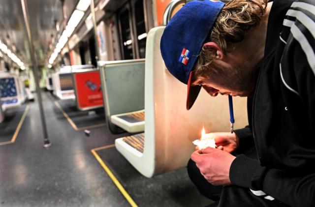At right, seated on a subway train, a young man with dark blonde hair and a baseball cap smokes fentanyl