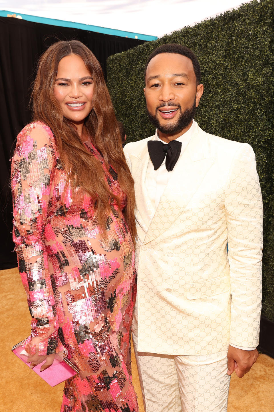 A pregnant Chrissy wears a sequined long-sleeved dress as she poses for a photo with John who's wearing suit and bowtie