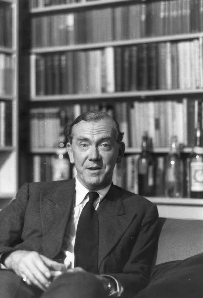 A man sits on a sofa in front of a bookshelf filled with books and bottles, wearing a suit and tie, holding a small object