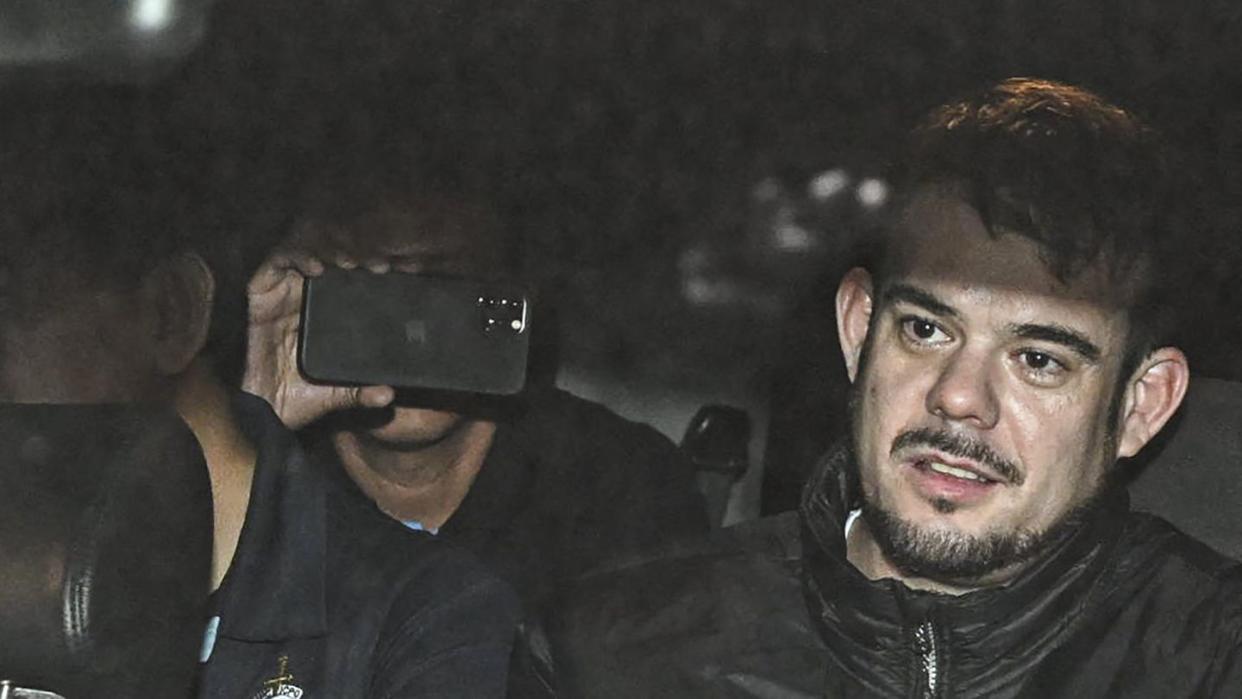 joran van der sloot sits in a car wearing a black zip up jacket, he looks past the camera with a neutral expression, two people sit on the left and are obscured by a seat headrest and a cell phone