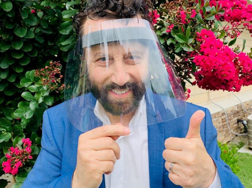 Yakov Smirnoff, smiling behind a face shield, gives a thumbs up