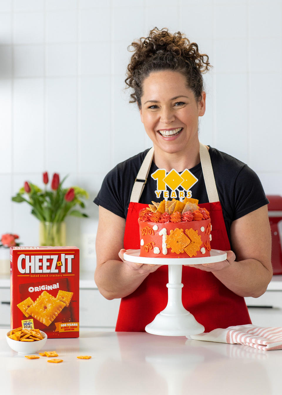 A chef holds a Cheez-It cake on a stand in a white room with a box of Cheez-it on the table
