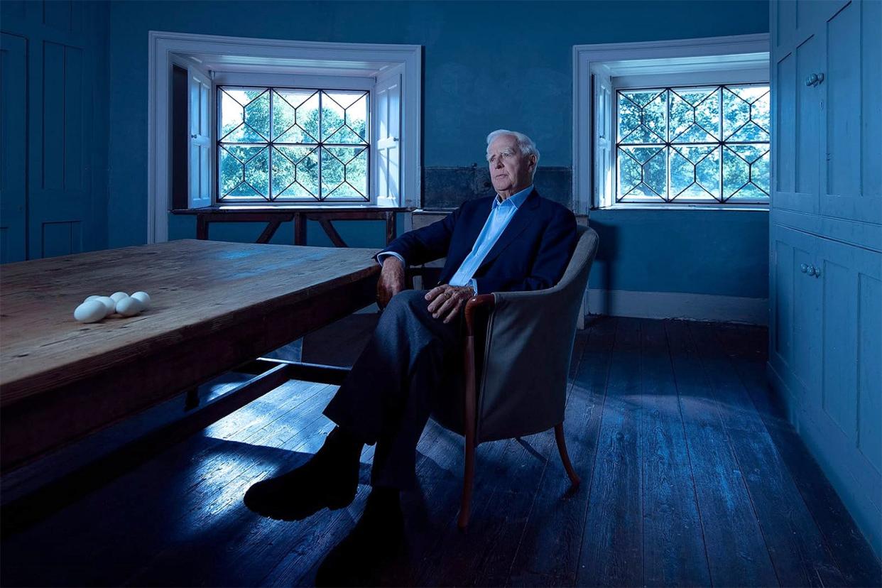 A older white-haired man, John le Carré, sits for an interview in a dimly lit room.