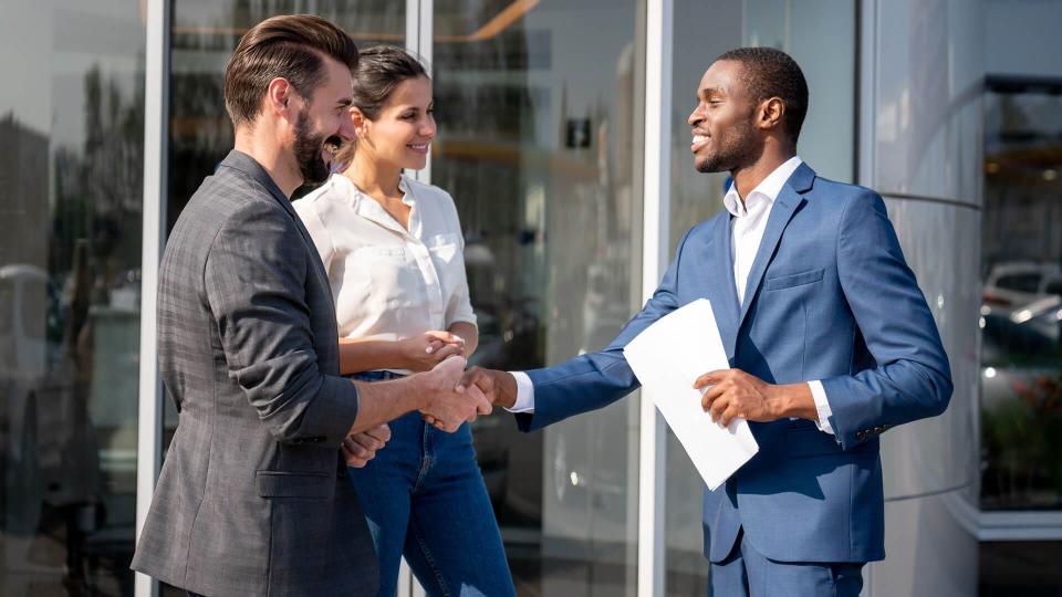 Portrait of a couple of happy customers closing a deal with a salesman at a car dealership and looking very happy handshaking.