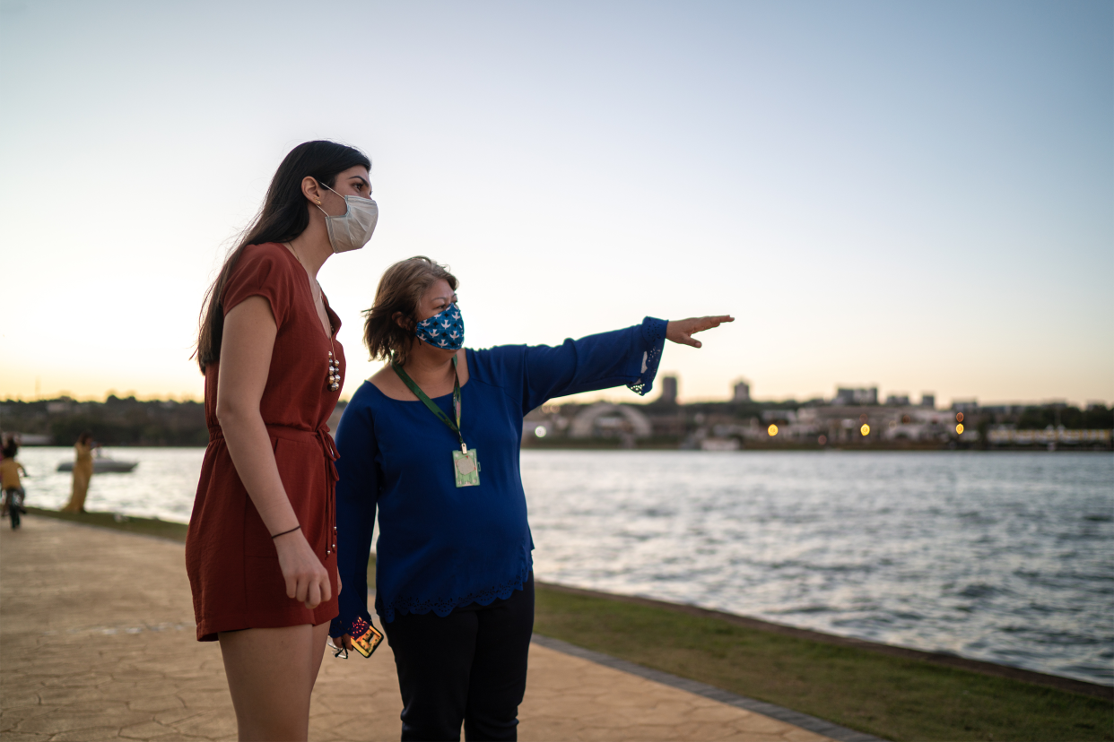 Senior woman pointing to the right for another woman with her to see, both wearing a face mask, on a sidewalk of a park with a river in the background, sun is setting