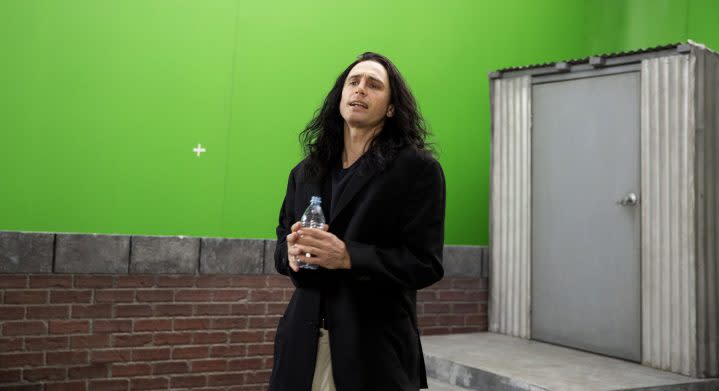 A man holds a water bottle and stands in front of a green screen.